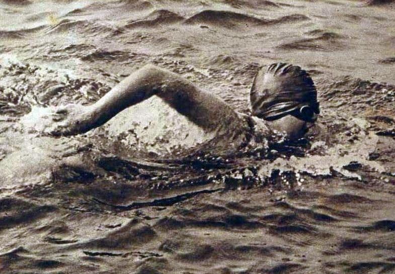 A swimmer wearing a period swim cap and goggles swimming freestyle in open water